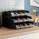 A Cambro Versa Black Self Serve 3-Tier Condiment Stand with 12in Bins filled with tea bags and sugar packets, placed on a kitchen counter.