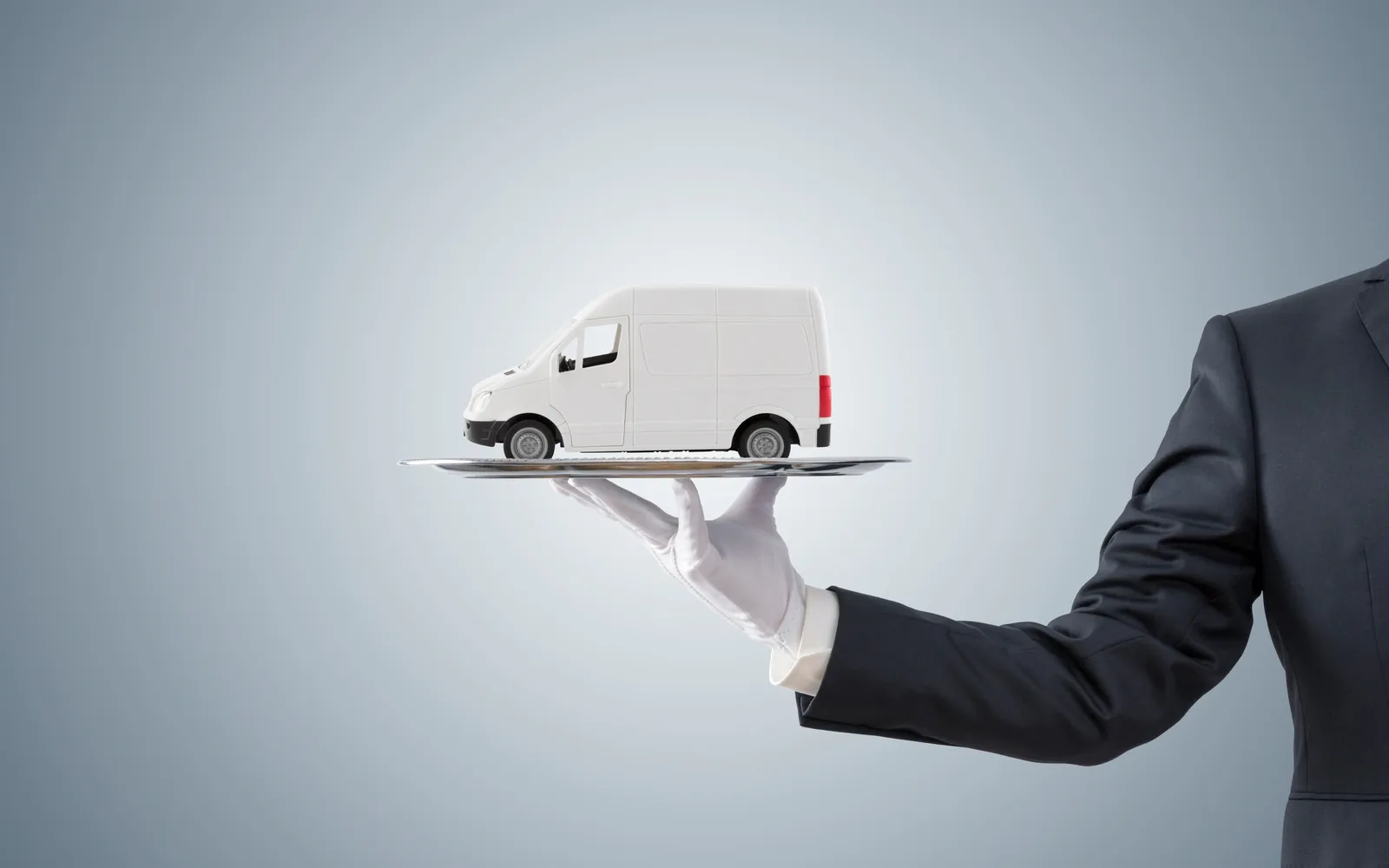 A person in a suit holding a small model of a white van on a flat palm, against a plain light blue background.