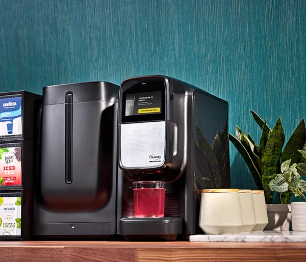 A Flavia® Chill Refresh for Creation 300 dispensing red liquid into a glass cup on a kitchen counter, with tea boxes and a plant beside it.