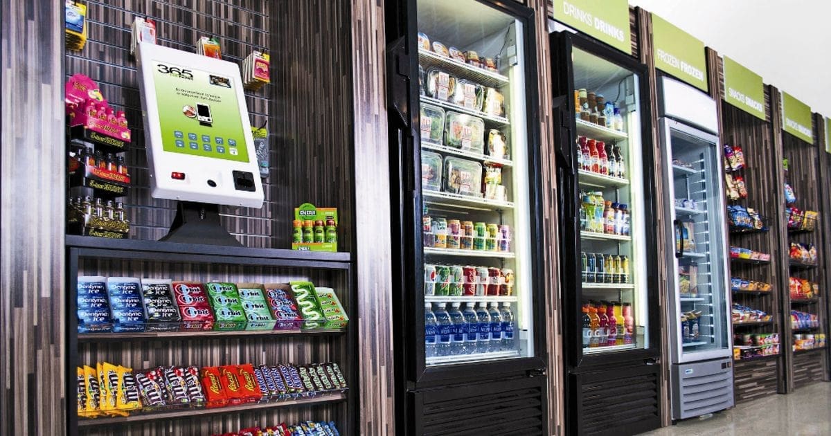 A modern vending area featuring an array of refrigerators filled with drinks and shelves stocked with snacks next to a digital payment terminal.